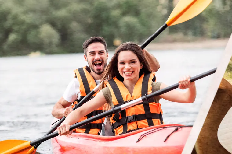 Pareja remando un kayak rojo con chalecos salvavidas, rodeados de árboles